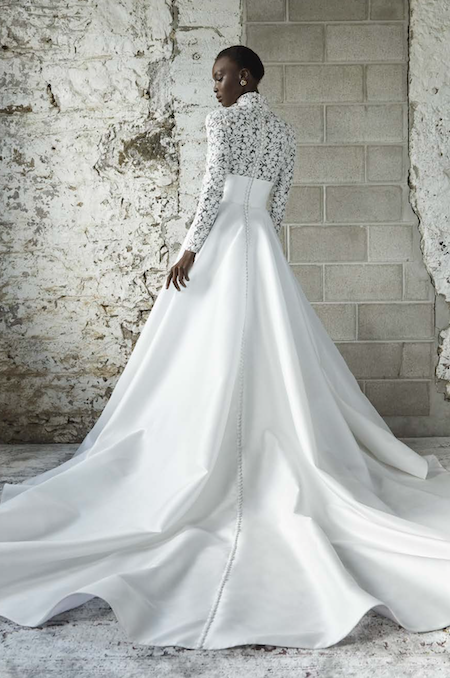 1738857282417-thyra-rivini2 Thyra, an ivory ballgown wedding dress by Rivini, featuring a detailed bodice with a refined mock neck and sculpted long sleeves adorned with intricate floral lace, a banded empire waist, and a full satin skirt with a chapel train accented by a row of covered buttons down the back, worn by a model looking over her shoulder against a textured wall.