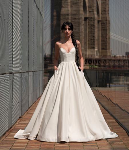 Crawford, an ivory satin ball gown by Alyne, features spaghetti straps and a V-neckline with a modified sweetheart cut. The full skirt includes pockets, and the chapel train extends onto a brick path. A model with a long dark braided ponytail poses between a textured wall and a reflective surface, with the Brooklyn Bridge visible in the sunlit background.