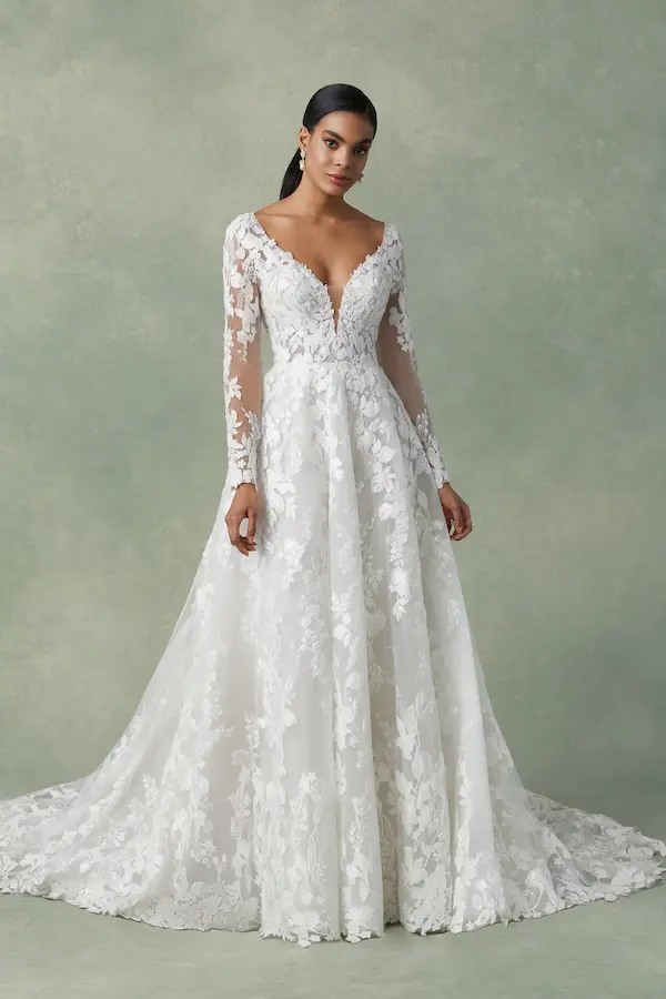 Freesia, a Sand/Ivory/Nude A-line bridal gown by Justin Alexander, features a plunging V-neckline, long illusion lace sleeves, delicate floral lace appliqués with beading, and a chapel length train.
