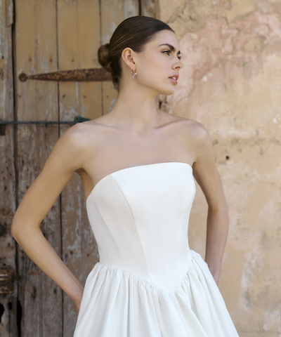 1738964138939-stcharles-annebarge St. Charles, a silk white Anne Barge strapless bridal ballgown, is modeled against a rustic wooden door and textured wall. The elegant gown features lustrous embroidered satin, a flattering gathered Basque waist, and a voluminous skirt, creating a sophisticated and graceful silhouette.