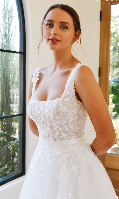 Ellie, an ivory ball gown bridal dress with a square neckline and tank straps, features a bodice adorned with delicate beading, lace, and floral appliques. A silk sash highlights the waist, leading to a full tulle skirt also embellished with matching lace and sequins. The model stands gracefully, looking directly at the viewer with a gentle smile, her hands clasped behind her back, next to a tall arched window.