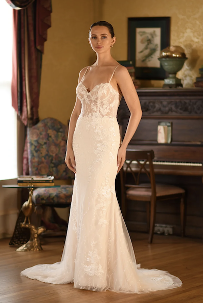 Lilith, an ivory and pearl mermaid wedding gown featuring a demi-sheer lace sweetheart bodice, delicate beaded spaghetti straps, and a chapel train adorned with sequins and lace appliques. A model poses in the elegant bridal dress within a warmly decorated room.
