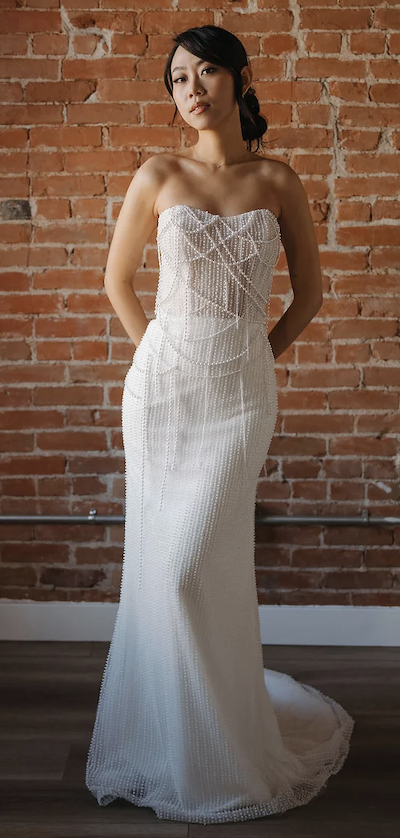 Monroe, an ivory fit-and-flare bridal gown by Sincerely Adorn, modeled by an Asian woman against a brick wall. The strapless dress features a sweetheart neckline, a bodice with intricate draping pearl beading, and a skirt adorned with vertical lines of beading that extend into a chapel train.