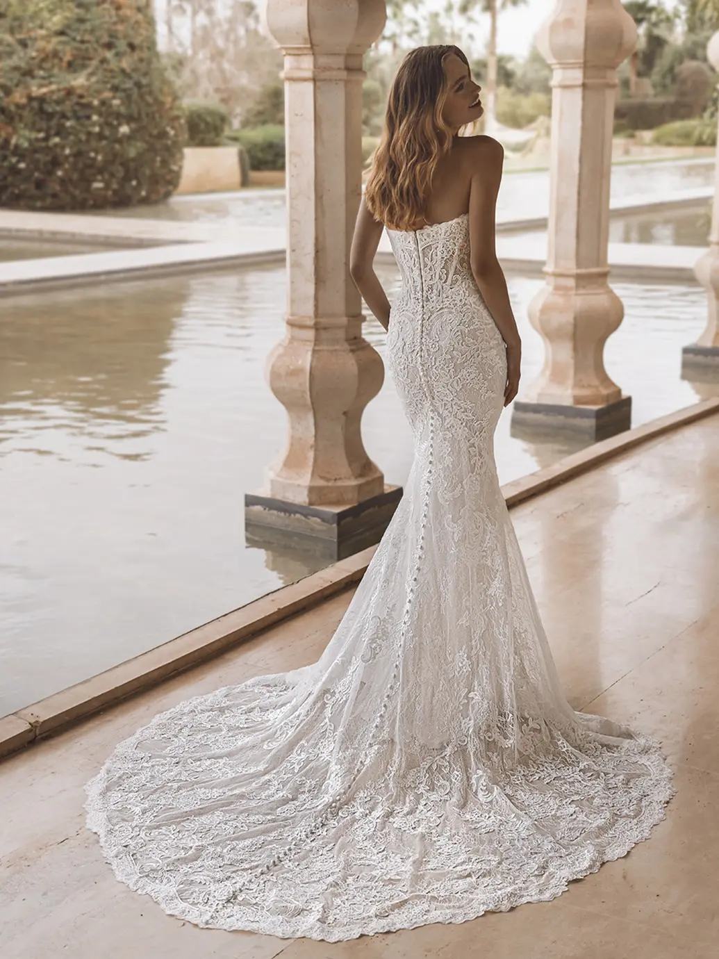1758907646166-26_Enzoani_PRO_VAIL_Bac_Web Vail, an ivory, sand, and nude fit-and-flare bridal gown by Enzoani, featuring delicate Alençon and Chantilly lace. This romantic strapless dress has a soft scoop neckline, a visible corset-style back, and a long row of buttons extending down its elegant cathedral train. The model, with wavy brown hair, stands with her back mostly to the viewer, looking over her shoulder at a tranquil water garden with classical columns.