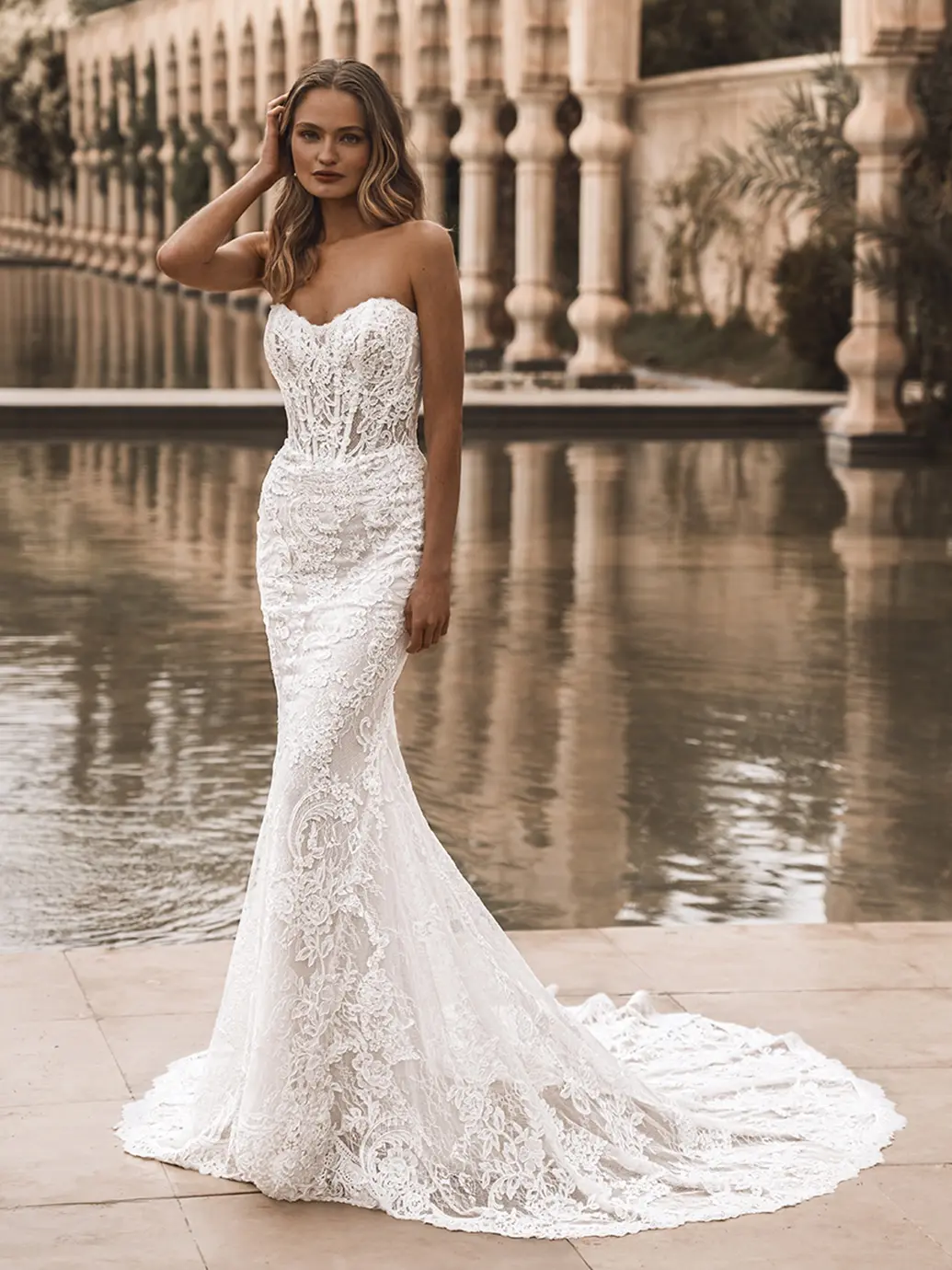 Vail, an Ivory, Sand, and Nude bridal gown, features a fit-n-flare silhouette with a strapless scoop neckline. The dress is intricately covered in lace, highlighting a corset-style bodice and adorned with delicate buttons. A long, flowing cathedral train trails behind. A model with wavy blonde hair, hand resting on her head, stands elegantly beside a serene reflective pool, with classical columns and architecture in the background.