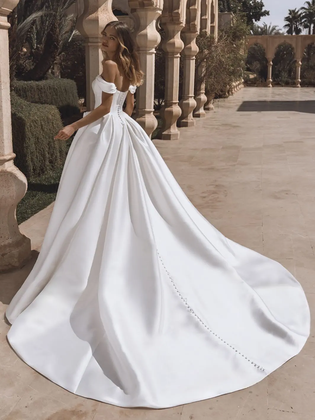 1758915057979-26_Enzoani_PRO_VIVIANA_Bac_Web_1064_1418_80_s_c1 Viviana, an ivory ballgown wedding gown crafted in rich mikado, features an off-the-shoulder scoop neckline. The back is adorned with a corset closure and a long line of delicate buttons cascading down its elegant cathedral train. The bride is captured from behind, looking over her shoulder at the viewer, walking on a paved pathway amidst architectural pillars and greenery.