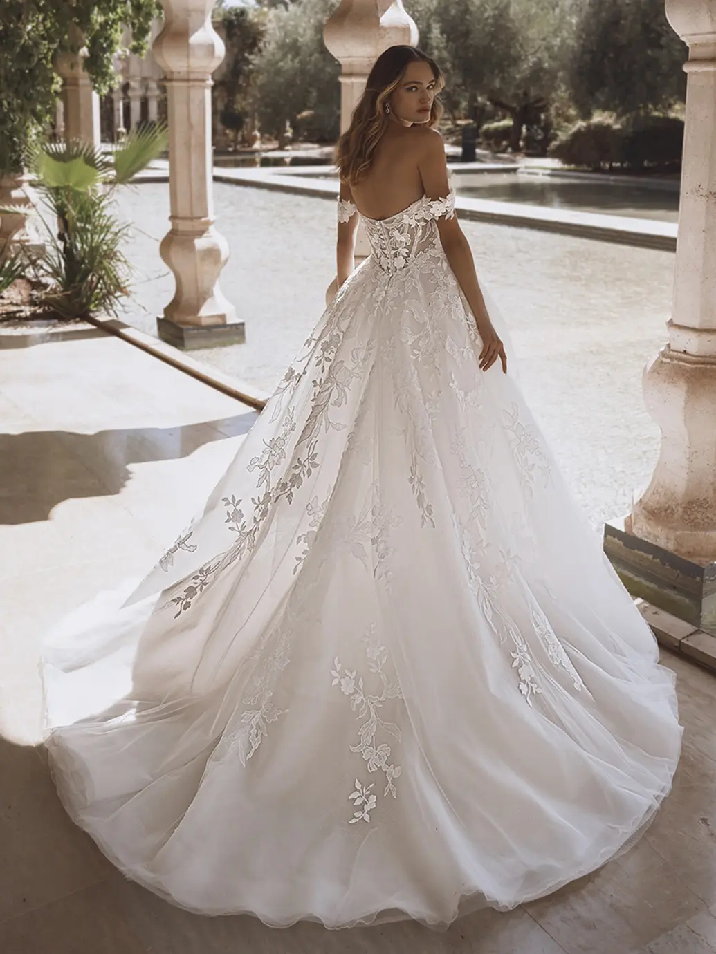 1758915273001-26_Enzoani_PRO_VOGUE_Bac_Web Vogue, an ivory, sand, and nude Enzoani ballgown, featuring a scoop neckline with detachable off-the-shoulder lace straps. The voluminous skirt of tulle and organza is adorned with delicate floral lace appliques that trail down to a long chapel train, visible from the corset and button-detailed back as the bride looks over her shoulder in an elegant outdoor garden.