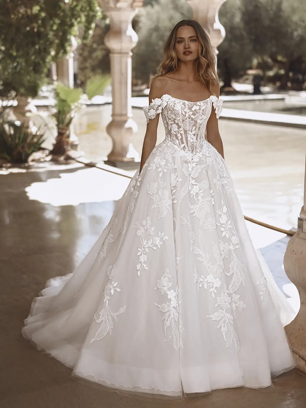 Vogue, an ivory, sand, and nude Enzoani ball gown wedding dress, features an off-the-shoulder design with an illusion corset bodice adorned with intricate lace appliques. These appliques gracefully extend onto the full tulle skirt, which has a basque waistline and a chapel train. The dress is modeled by a woman with long wavy hair, posing in a sunny outdoor setting with classical architectural elements, columns, and a serene water feature.