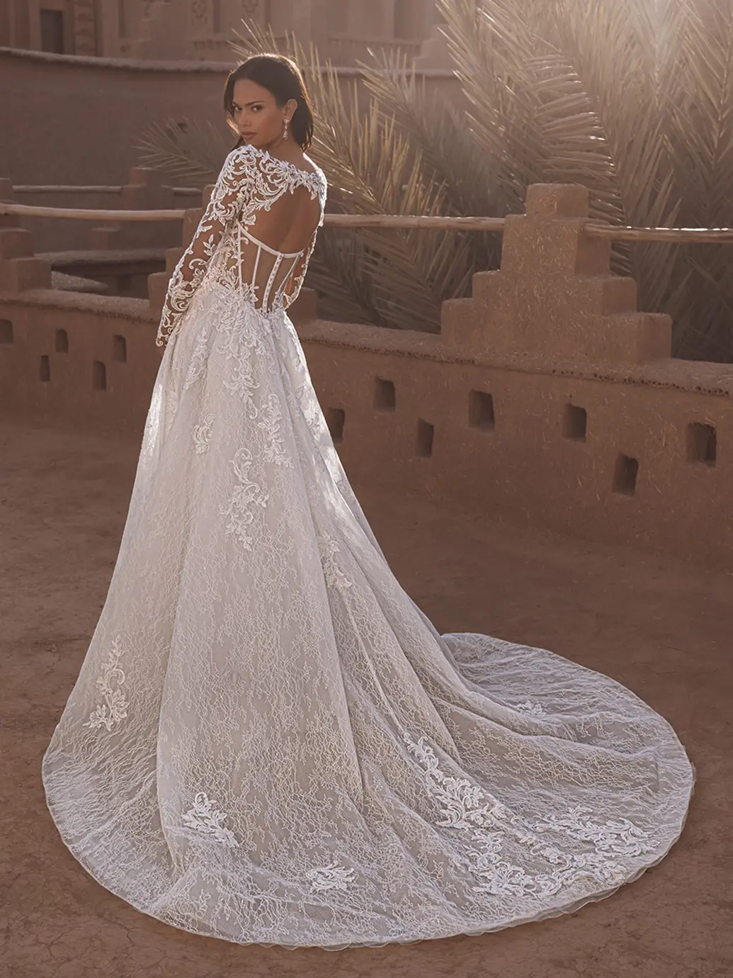 1758915799388-26_blue_pro_tabitha_bac_web Tabitha, an ivory and nude A-line wedding gown by Blue by Enzoani, crafted from delicate Alençon lace with intricate appliques. It features long lace sleeves, a soft scoop neckline, and a captivating keyhole back with a structured corset and exposed boning. The chapel-length train flows behind the model, who looks over her shoulder in an outdoor setting with warm adobe walls.