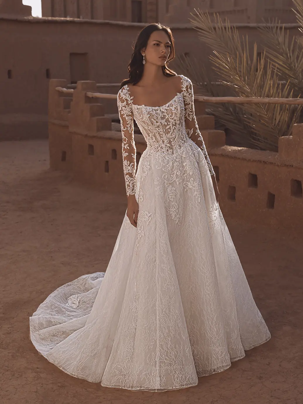 Tabitha, an ivory and nude A-line bridal gown featuring a structured, illusion bodice adorned with intricate lace appliqués, long illusion sleeves, a scoop neckline, and a chapel-length train.