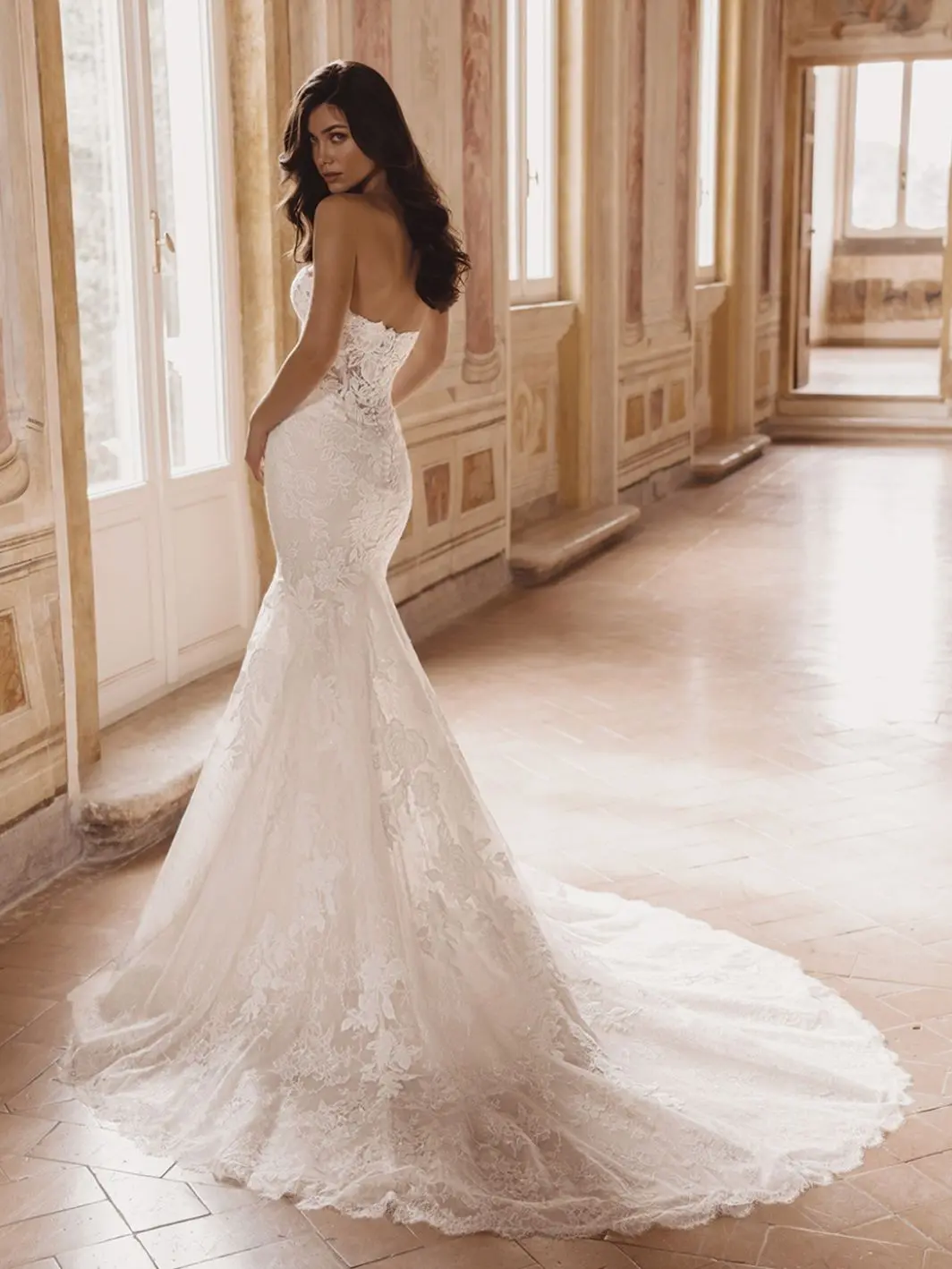 1758916437771-26_Blue_PRO_TATE_Bac_ColorEdit_Web_1064_1418_80_s_c1 Tate, an ivory and nude mermaid wedding gown crafted from delicate floral embroidered lace and Chantilly lace, featuring a strapless sweetheart neckline. The fitted silhouette boasts intricate lace detailing throughout and extends into a dramatic chapel train, worn by a model in a sunlit classical hallway.