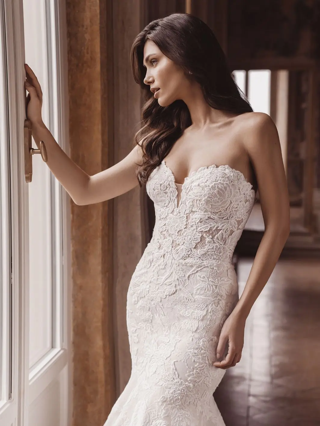 1758916437771-26_Blue_PRO_TATE_Clo_ColorEdit_Web_1064_1418_80_s_c1 Tate, an ivory and nude mermaid wedding gown crafted from ethereal floral embroidered and Chantilly lace, features a strapless sweetheart neckline and a sculpted, couture-inspired finish. The gown is worn by a model with long, wavy dark hair, who stands elegantly looking out a window with one hand resting on the window frame.