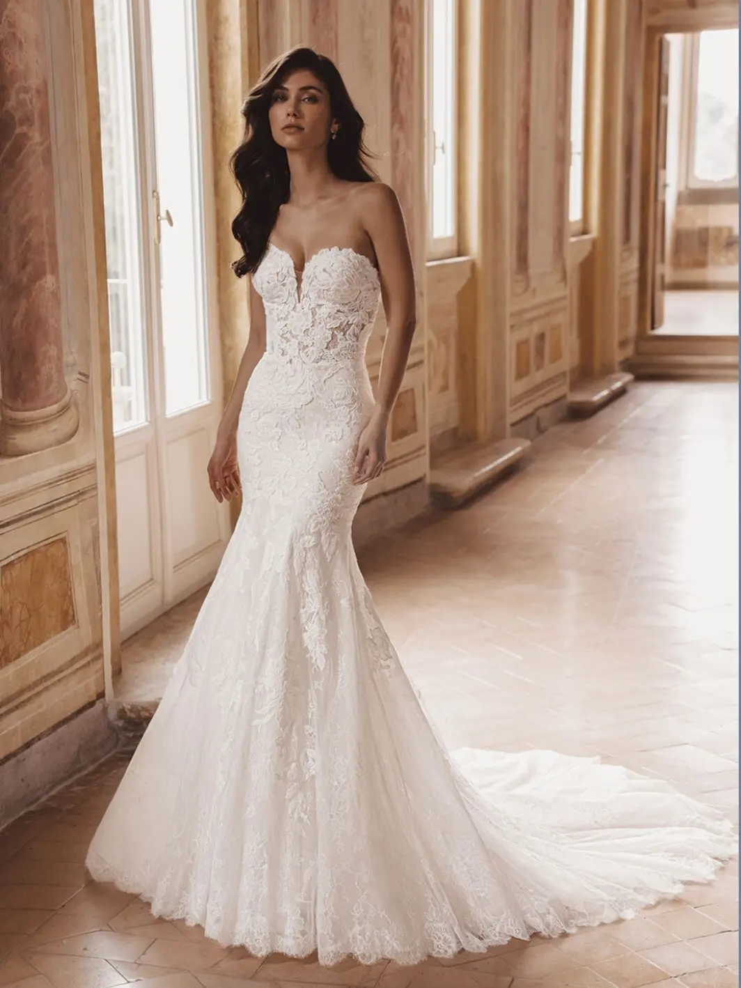 Tate, an ivory and nude mermaid bridal gown featuring a strapless sweetheart neckline, delicate floral lace, and an illusion corset bodice. The fitted silhouette extends into a chapel-length train with a scalloped lace hem. A model with long dark hair poses in the elegant gown within a grand hall.