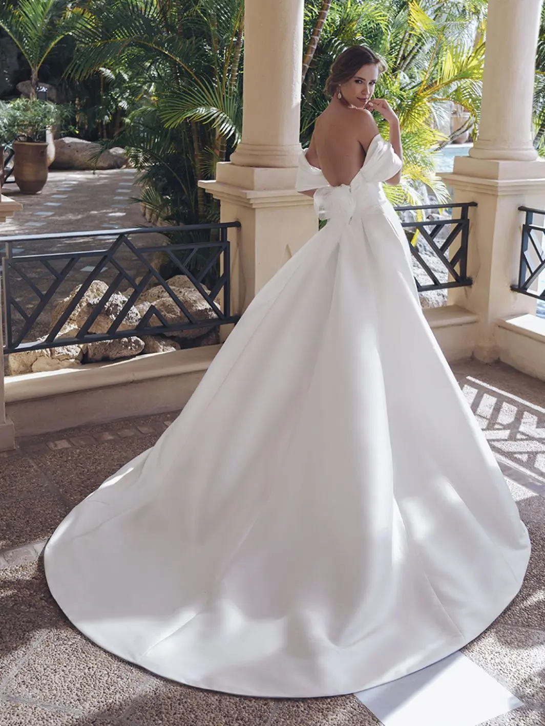 1758917666872-25-blue-pro-sevilla-sleeves-bac-web-1064-1418-80-s-c1 Sevilla, an ivory A-line wedding gown crafted in luxurious Mikado, features delicately draped, detachable off-the-shoulder straps, an open corset back with a tulle bow, and an elegant draped bodice. The pleated skirt boasts hidden pockets and extends into a voluminous cathedral train. A bride models the gown from behind, looking over her shoulder at the viewer, on a patio surrounded by tropical foliage and pillars.
