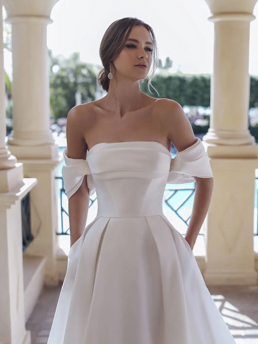 1758917666872-25_Blue_PRO_SEVILLA_Sleeves_Clo_Web_1064_1418_80_s_c1 Sevilla, an ivory A-line wedding gown, featuring a draped bodice with off-the-shoulder straps, and a voluminous pleated Mikado skirt with pockets. A model with an elegant updo poses wearing the gown in a sunlit outdoor setting with classical pillars.