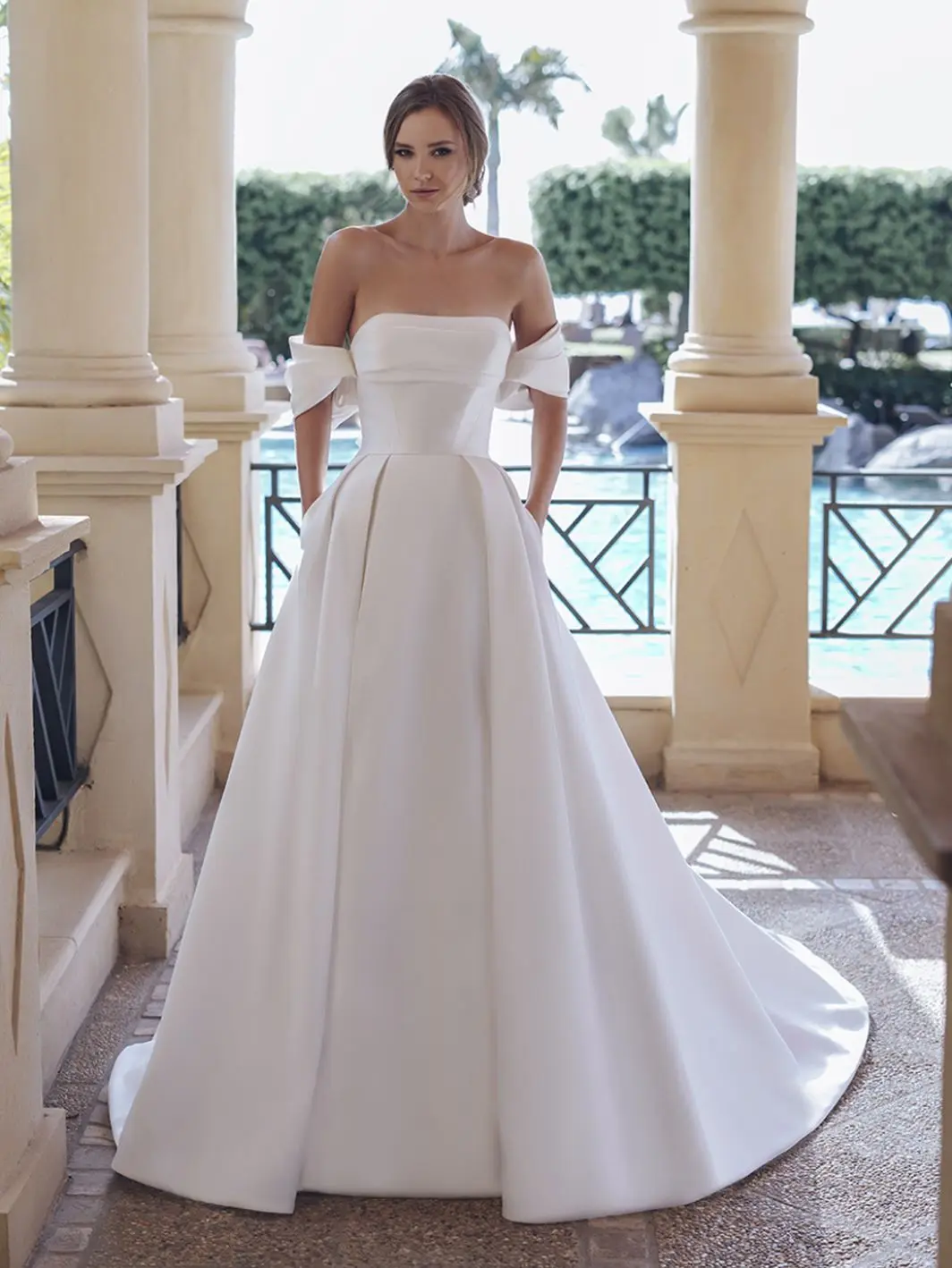 1758917666873-25-blue-pro-sevilla-sleeves-fro-web-1064-1418-80-s-c1 Sevilla, an ivory A-line bridal gown featuring a strapless, draped bodice with off-the-shoulder bow details, and a voluminous pleated Mikado skirt with pockets, flowing into a cathedral train. A woman with an elegant bun models the gown on a stone patio overlooking a pool.