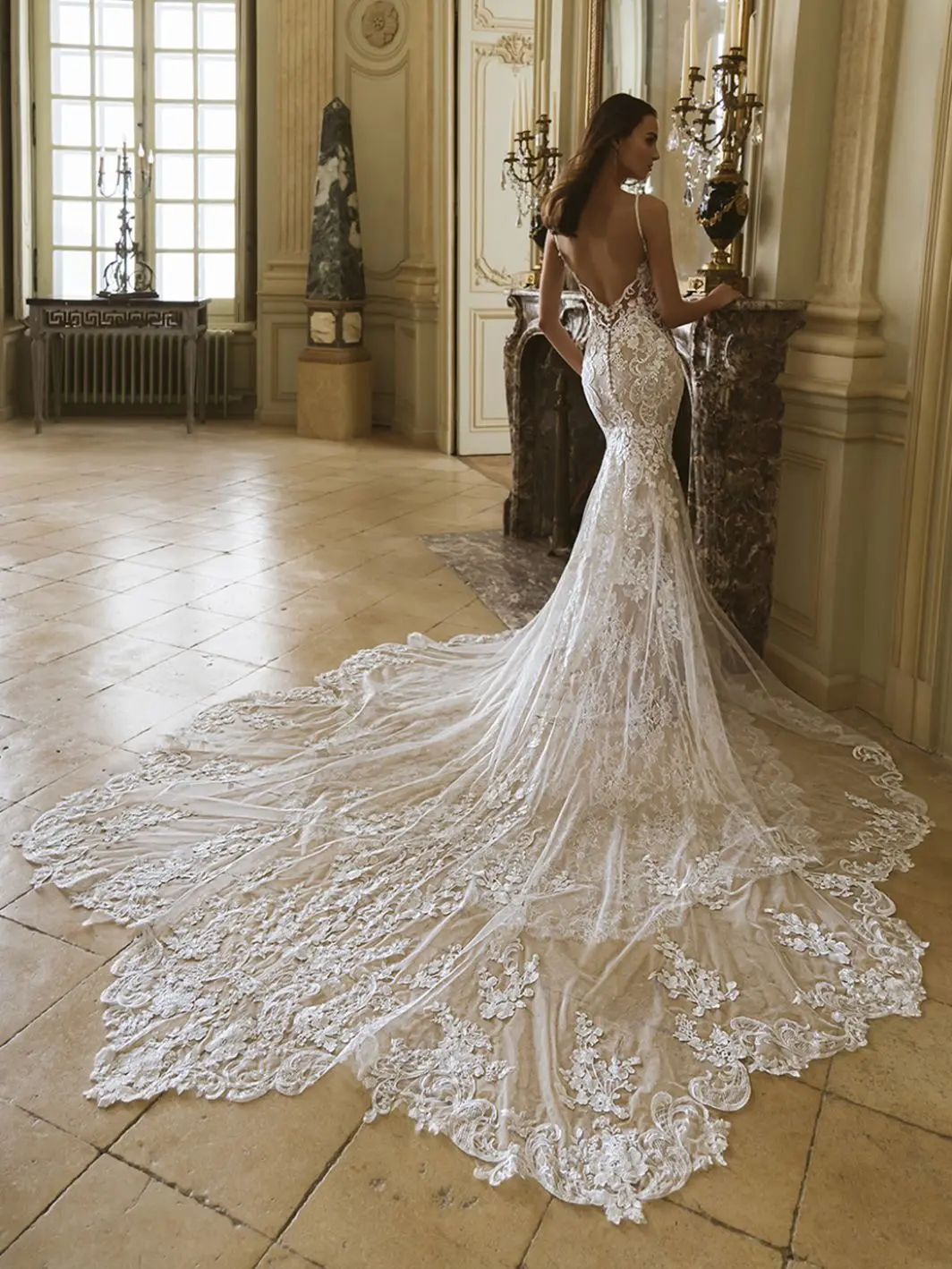 1759505904043-26_Elysee_PRO_GEM_Bac_Web_1064_1418_80_s_c1 Gem, an ivory and nude mermaid wedding gown from the Élysée collection, features ethereal baroque lace with lush floral embroidery and a dramatic cathedral train. The dress has delicate spaghetti straps, a plunging V-neckline, an illusion bodice with buttons down the open back, and intricate beading. A model with long dark hair stands with her back to the viewer, looking over her shoulder, in an opulent room with ornate fireplace, candelabras, and large windows.