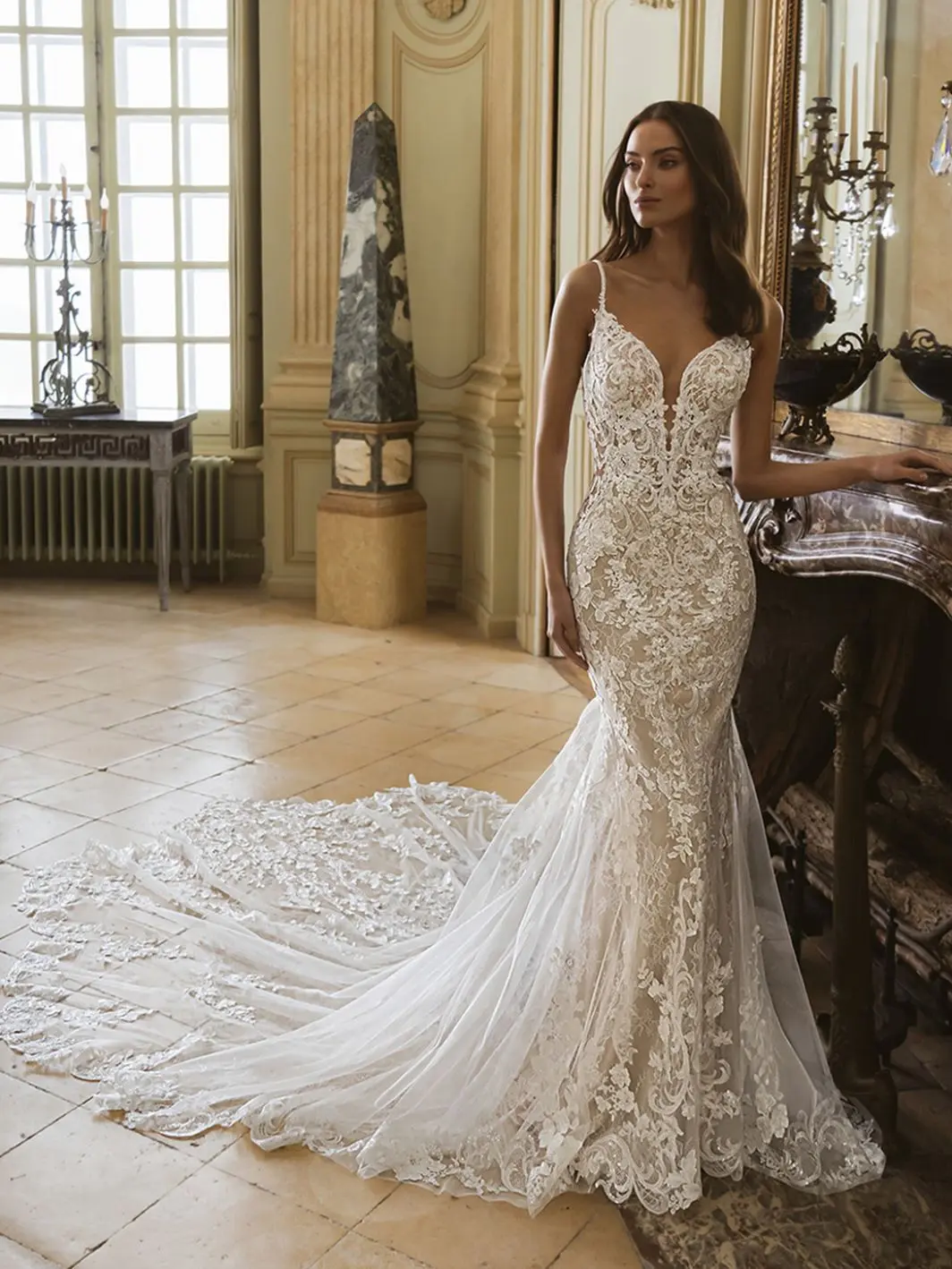 1759505904044-26_Elysee_PRO_GEM_Fro_Web_1064_1418_80_s_c1-1 Gem, an Ivory/Nude/Nude mermaid wedding gown featuring delicate lace with intricate floral embroidery and subtle beading. The dress has a plunging V-neckline with illusion details, thin spaghetti straps, an open back, and buttons running down the back. A dramatic cathedral-length train, adorned with scattered floral appliques, trails elegantly on the tiled floor. The model stands gracefully in a grand, classically decorated room.