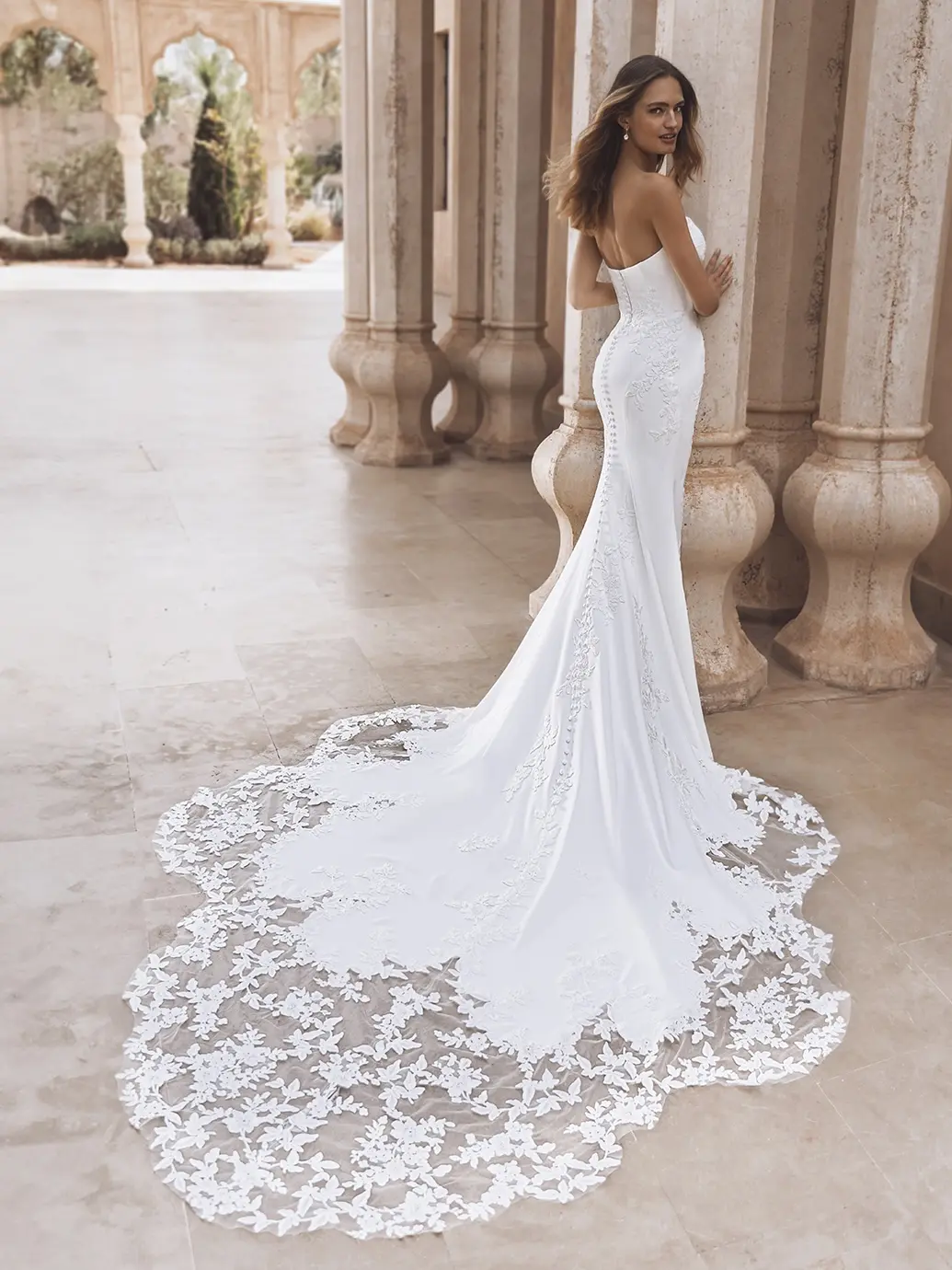 1760459449068-uploads-1758909640153-26_Enzoani_PRO_VEDA_Bac_Web Veda, an ivory fit-and-flare bridal gown by Enzoani, features a strapless scoop neckline and a sleek crepe silhouette with delicate floral embroidered lace on the bodice and a stunning button-back. A breathtaking cathedral-length train, intricately embellished with floral lace cutouts, trails gracefully behind the bride as she poses elegantly against stone pillars.