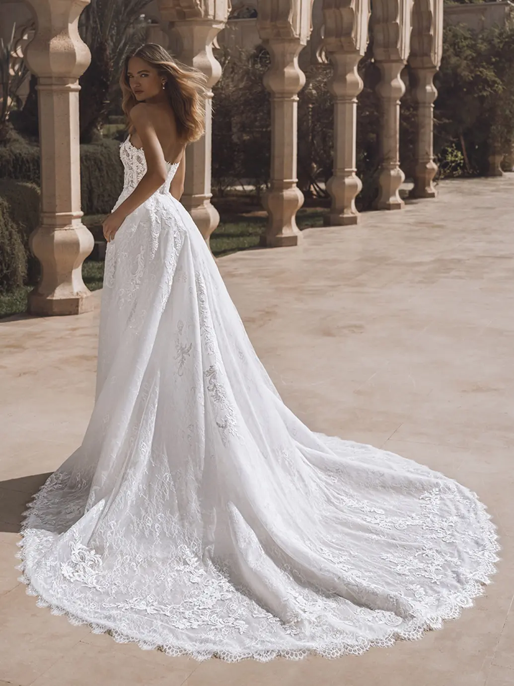 1760459598589-26_Enzoani_PRO_VENICE_Bac_Web Venice, an ivory and nude Enzoani bridal gown, is a vintage-inspired strapless A-line masterpiece. Crafted from pearl-beaded floral and corded lace, it features a detachable overskirt flowing into an elaborate cathedral train. The gown is worn by a model looking back over her shoulder, posed outdoors against an ornate columned backdrop.