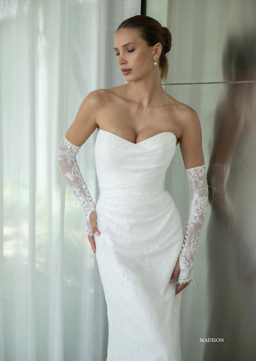 1762533873795-screenshot-2025-11-07-at-10-43-36-am Madison, an ivory sheath-style fit-and-flare bridal gown crafted from delicate lace. It features a strapless, modified sweetheart neckline, a draped bodice that flatters the natural waist, and a softly pleated skirt, finishing with a chapel-length train. The model is elegantly posed, wearing matching fingerless lace gloves.