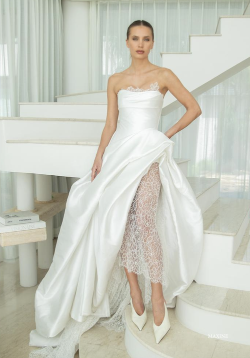 Maxine, a white Mariana Hardwick bridal gown featuring a structured, hand-draped strapless bodice with delicate lace embellishments. Its dramatic A-line Mikado skirt is elegantly draped, revealing a full lace underskirt and a chapel-length train. The gown is worn by a model standing on white marble stairs in a brightly lit, modern room.
