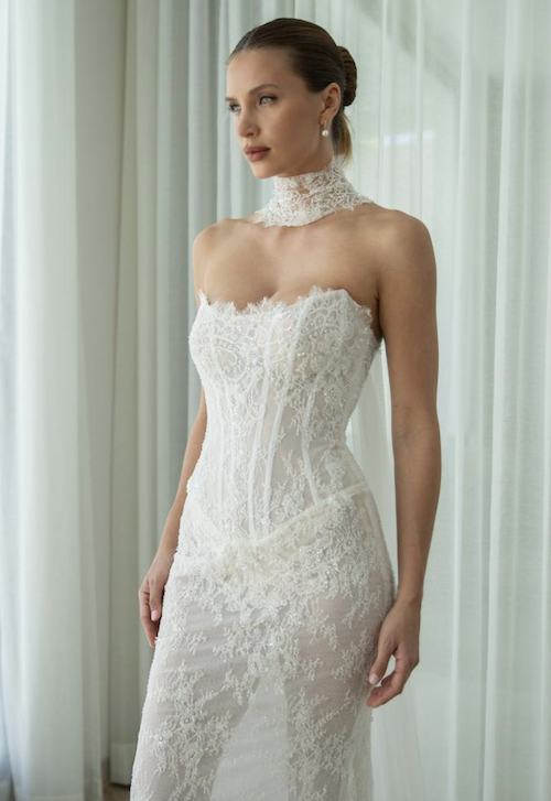 1762538211826-screenshot-2025-11-07-at-11.55.47-am Iris, an ivory and nude fit-and-flare bridal gown, showcasing sculptural, form-fitting lace with delicate beading over a structured, strapless corset bodice with a scoop neckline and visible boning. The gown is paired with a matching detachable lace choker. The model looks thoughtfully to the side against a soft, light background.