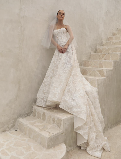 Delilah, an ivory and nude A-line bridal gown by Mariana Hardwick, features a strapless structured corset bodice with a scoop neckline and delicate lace detailing. The dress has a sweeping cathedral train. A model in a short veil stands on a light stone staircase, leaning against a textured wall.