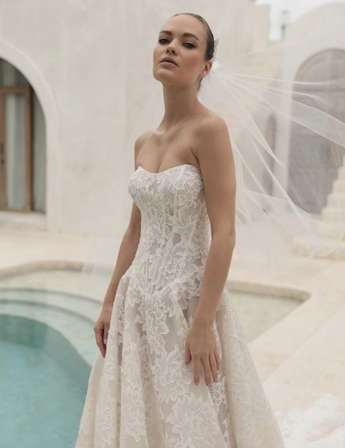 1762538928626-screenshot-2025-11-07-at-12-07-21-pm Delilah, a romantic ivory lace A-line wedding gown with a nude lining, featuring a strapless scoop neckline and a structured corset bodice with a defined Basque waist. The full lace skirt extends into a sweeping cathedral-length train. The model is posed outdoors next to a pool, her veil flowing in the breeze, with white architectural elements in the background.