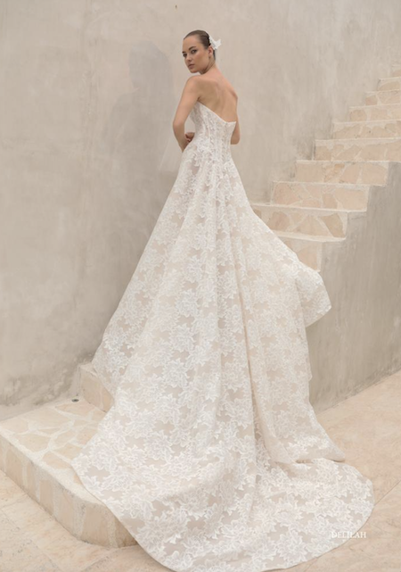 1762538928627-screenshot-2025-11-07-at-12-07-33-pm Delilah, a romantic A-line wedding gown by Mariana Hardwick, features off-white lace over a nude lining. It has a strapless scoop neckline, a structured corset bodice with a Basque waist, and an alluring open back. The full lace skirt flows into a sweeping cathedral-length train. The dress is modeled by a woman with her hair pulled back and a white flower, standing on stone steps and looking over her shoulder.