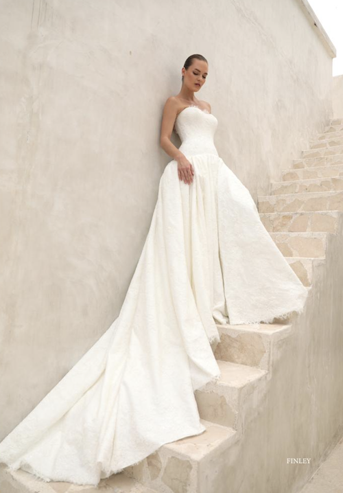 Finley, an ivory A-line bridal gown by Mariana Hardwick, featuring a dropped waist, strapless lace bodice, open back, button detailing, and a sweeping cathedral train.