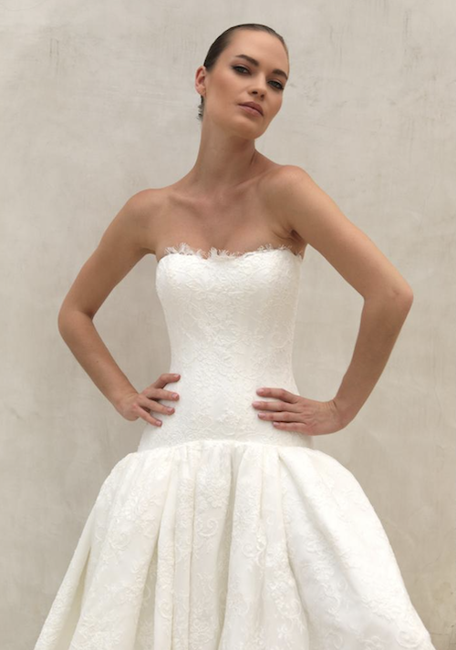 1762539047698-screenshot-2025-11-07-at-12-09-48-pm Finley, an ivory A-line wedding gown by Mariana Hardwick. The strapless dress features a fitted lace bodice, a dropped waist, and a voluminous skirt. A model with hands on her hips poses confidently against a light-colored wall.