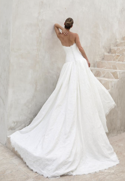 1762539047698-screenshot-2025-11-07-at-12-09-54-pm Finley, an ivory A-line wedding gown with a strapless bodice, dropped waist, and an open back with a line of buttons, featuring a flowing lace skirt and a sweeping cathedral-length train. The model is shown from behind, leaning against a textured wall next to a stone staircase.
