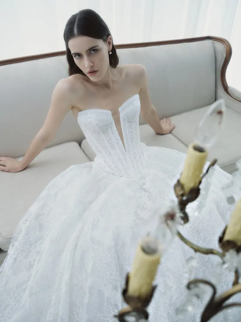 Radiance, an ivory Rivini bridal ball gown, featuring a strapless deep V-neck corset bodice with a basque waist, and a full skirt adorned with intricate lace and tulle applique. A model with dark hair is seated on a light sofa, gazing towards the viewer, with a vintage chandelier partially in the foreground.