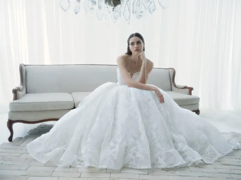 1766172671518-app8plprevyay3n-d-5505-radiance-fw26-rv-rivini-web-1 Radiance, an ivory Basque waist ballgown wedding dress crafted in delicate Chantilly corded lace, features a deep V-neck corset bodice softly veiled in draped tulle and adorned with hand-appliquéd lace, with a graceful line of buttons trailing to the hem of its expansive skirt. A model wearing the strapless gown sits with a thoughtful expression on a white sofa in a bright room with sheer white curtains and a chandelier.