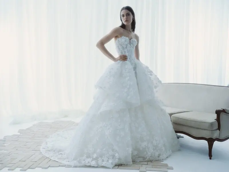 Reverie, an ivory Rivini ball gown wedding dress, features a structured, strapless corset bodice with a modified sweetheart neckline, intricate 3D floral lace, and beading. Its dramatic, voluminous skirt is tiered organza and lace, adorned with 3D floral appliqués, pickups, and a chapel train. A model with dark hair poses with her hand on her hip, looking forward, in a bright studio with sheer white curtains, a white patterned floor, and a white vintage sofa to the right.