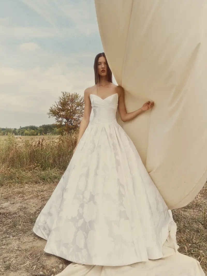 Velour, an ivory bridal ball gown by Alyne, featuring a strapless V-neck, a draped bodice, and a full pleated skirt in a floral jacquard fabric with a cathedral train. A model wears the gown in an outdoor field, holding a large, light-colored fabric prop.