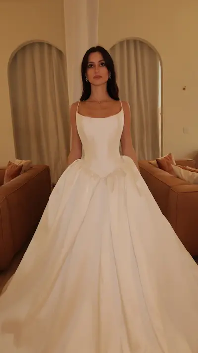 Adella, an ivory bridal gown by Sincerely Adorn, features an A-line silhouette with delicate spaghetti straps, a square neckline, and a flattering basque waistline. Crafted from lustrous Mikado fabric, the gown showcases a beautifully pleated skirt that flows into a chapel-length train. A woman with dark hair models the dress, standing elegantly in a well-lit room with arched architectural details and warm-toned furniture in the background.
