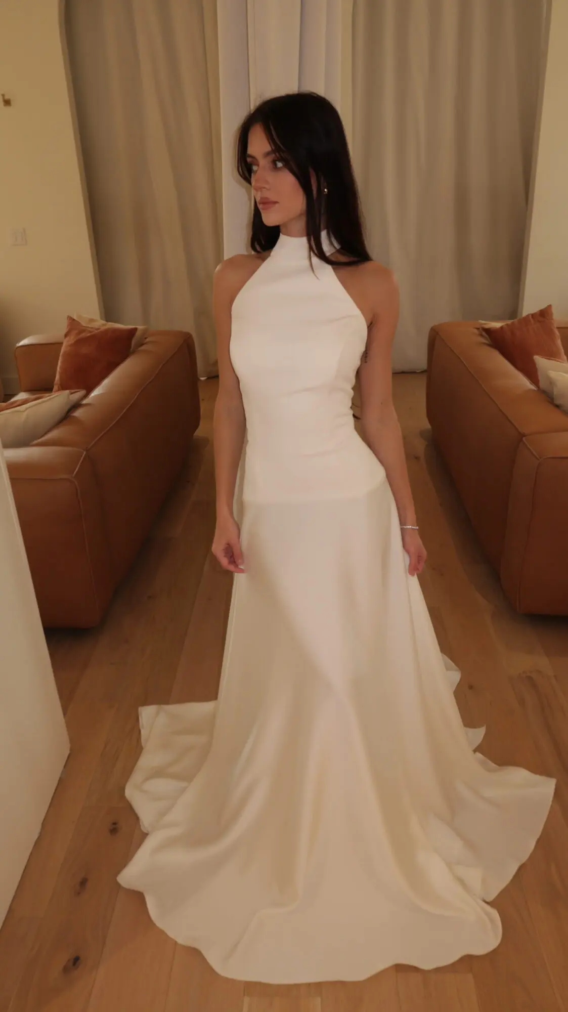 A woman models the Monroe bridal gown by Sincerely Adorn. This ivory dress features a 2-piece look with a high halter neckline and a dropped waist. The slim A-line silhouette is crafted from charmeuse and chiffon, flowing to a floor-length skirt with a train.