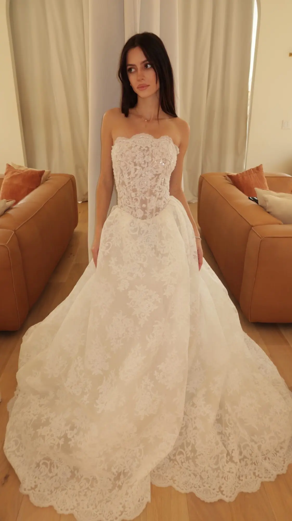 Valerie, an ivory and nude A-line bridal gown by Sincerely Adorn, featuring a strapless demi-sheer lace corset bodice and a full lace skirt with a chapel train. A woman with long dark hair models the dress indoors, looking to her left, with tan leather couches and light curtains in the background.