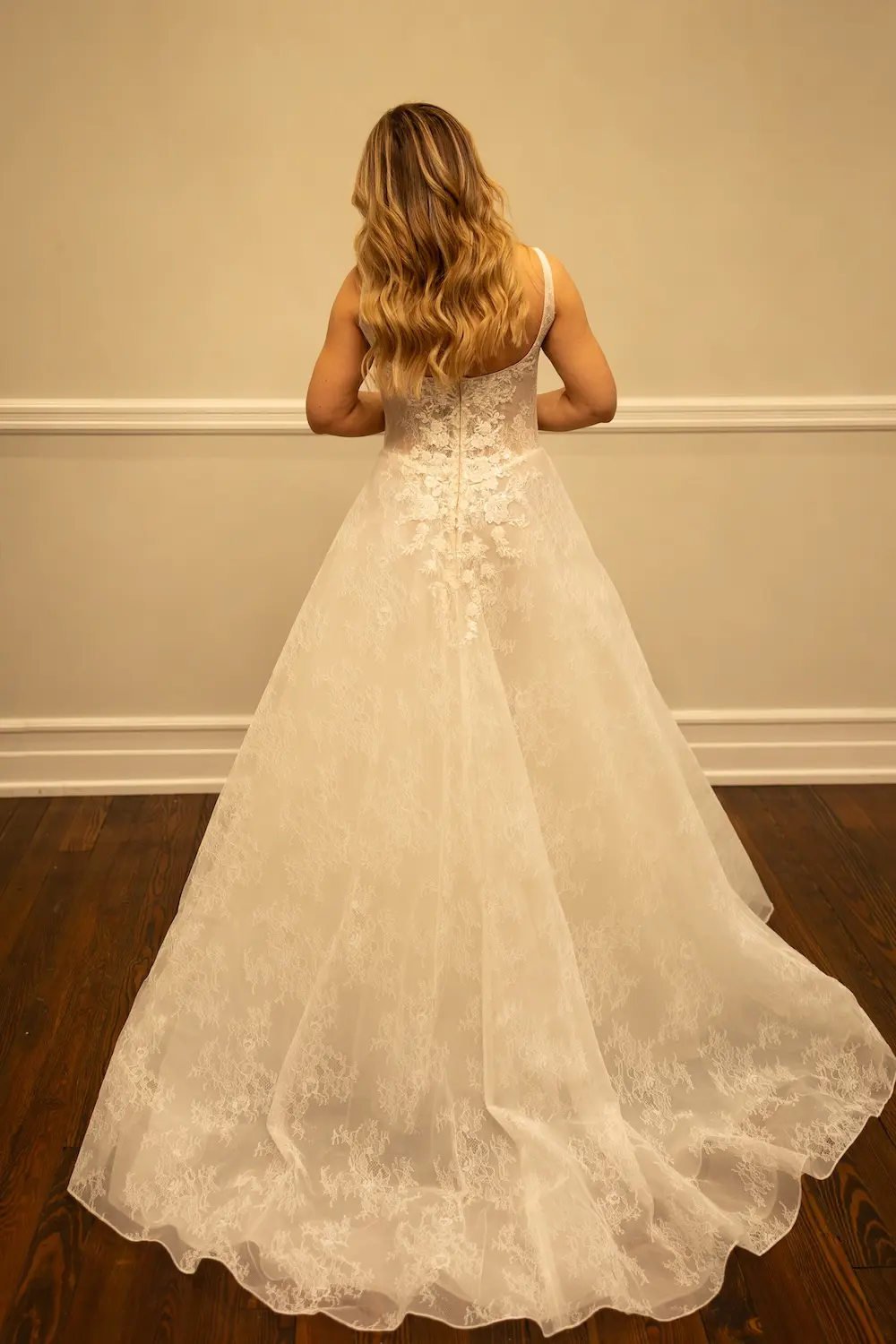 1771346001394-RH006 Savanna, an ivory A-line wedding gown with delicate spaghetti straps, is seen from the back. The romantic dress features a demi-sheer bodice with a flattering Basque waist and shimmering beaded lace appliqués that adorn the gown from top to its airy, chapel-length train, flowing gracefully on a dark wooden floor.