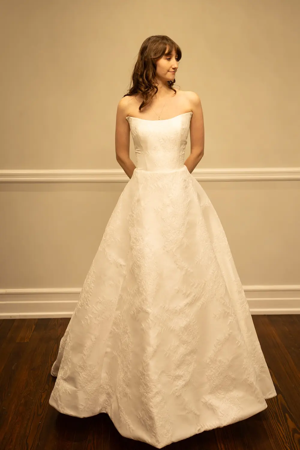 Vivienne, an ivory A-line bridal gown by Sincerely Adorn, features a strapless scoop neckline, a natural waist, and a chapel train, crafted from beautiful jacquard fabric. A woman with long wavy brown hair models the full-length dress, standing with her hands clasped behind her back, looking to the side against a light-colored wall and dark wooden floor.