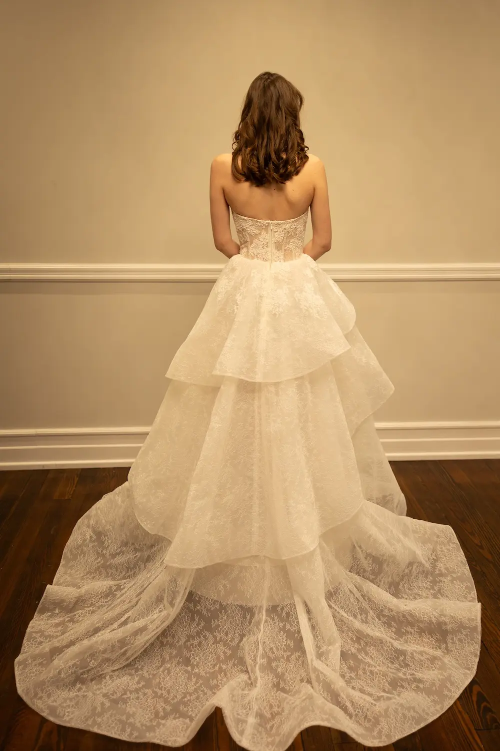 1771346958581-rh030-1 Florence, an ivory and nude A-line wedding gown with a strapless, demi-sheer lace bodice featuring exposed boning, a dramatic asymmetrical ruffled skirt, and a long lace train on the floor. The gown is shown from the back.