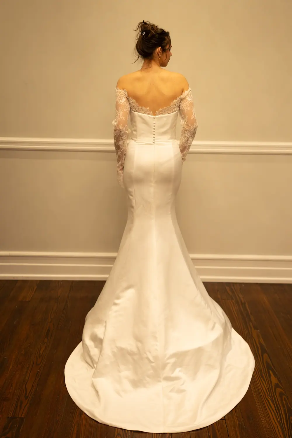 1771348036866-rh005-1 Madison, an ivory satin mermaid wedding gown, is seen from the back. It features a strapless neckline with delicate off-the-shoulder lace sleeves and a line of covered buttons down the back. The gown hugs the body through the hips before flaring into a dramatic chapel-length train.
