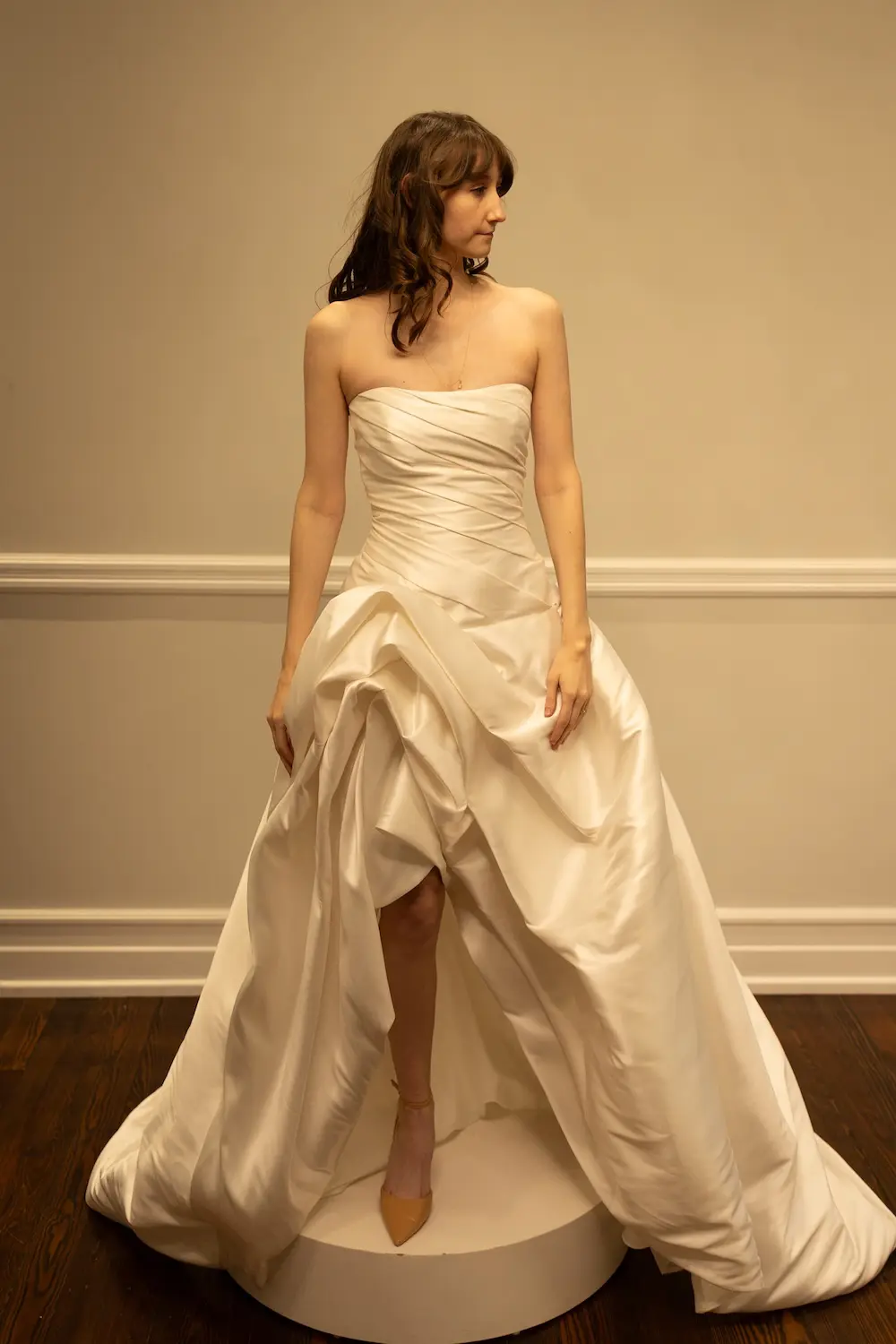 Blair, a Diamond White Mikado strapless A-line wedding gown by Sincerely Adorn, featuring a draped bodice, dropped waist, and a dramatic asymmetric draped skirt with a chapel train that reveals one leg. The model has long wavy hair and wears nude heels.