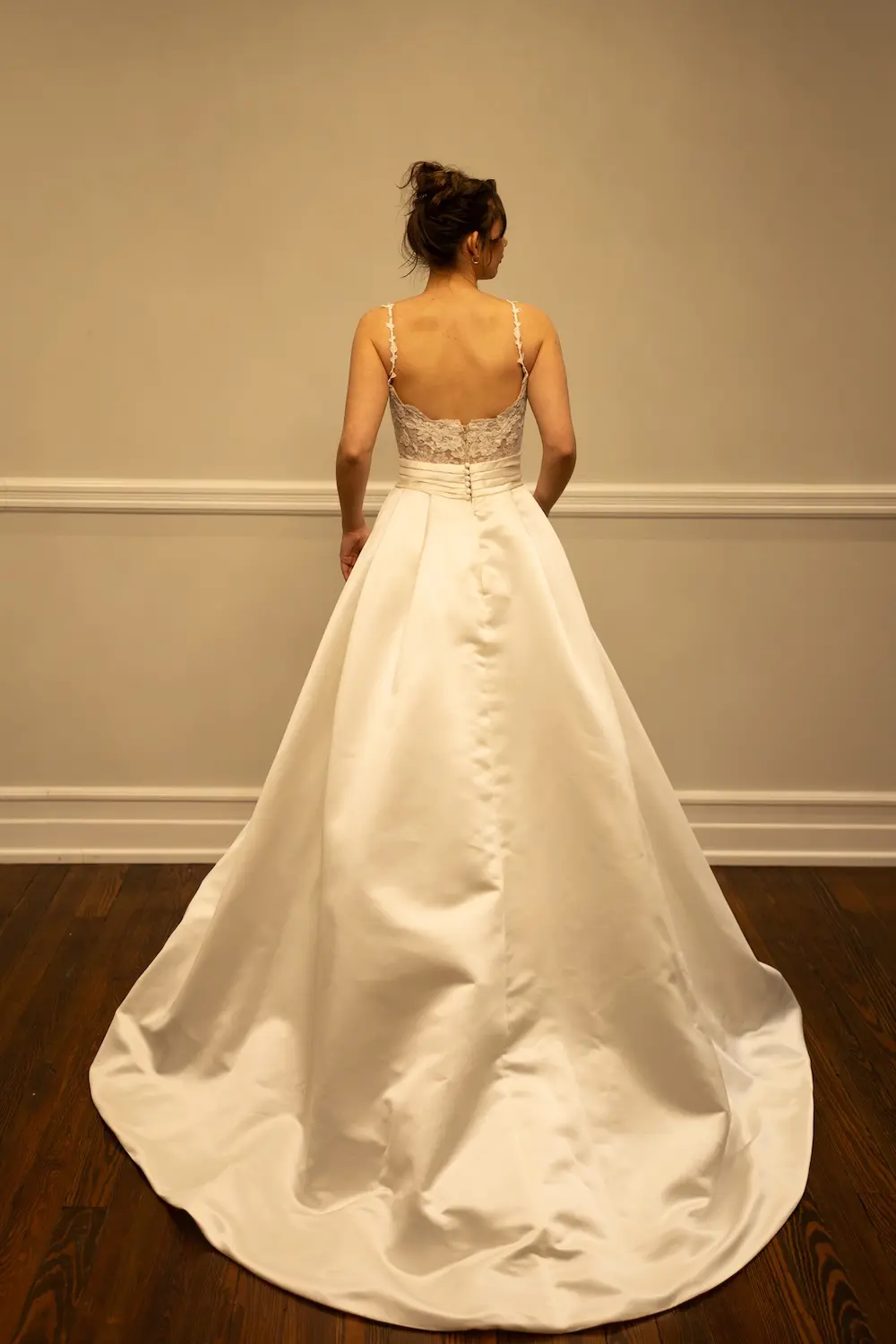 1771349064770-RH023 Morgan, an ivory/nude ballgown wedding dress featuring a semi-sheer lace bodice with delicate buttons down the back, lace spaghetti straps, and a soft scoop neckline, is shown from behind on a model. The lustrous satin skirt flows into a sweeping chapel-length train.