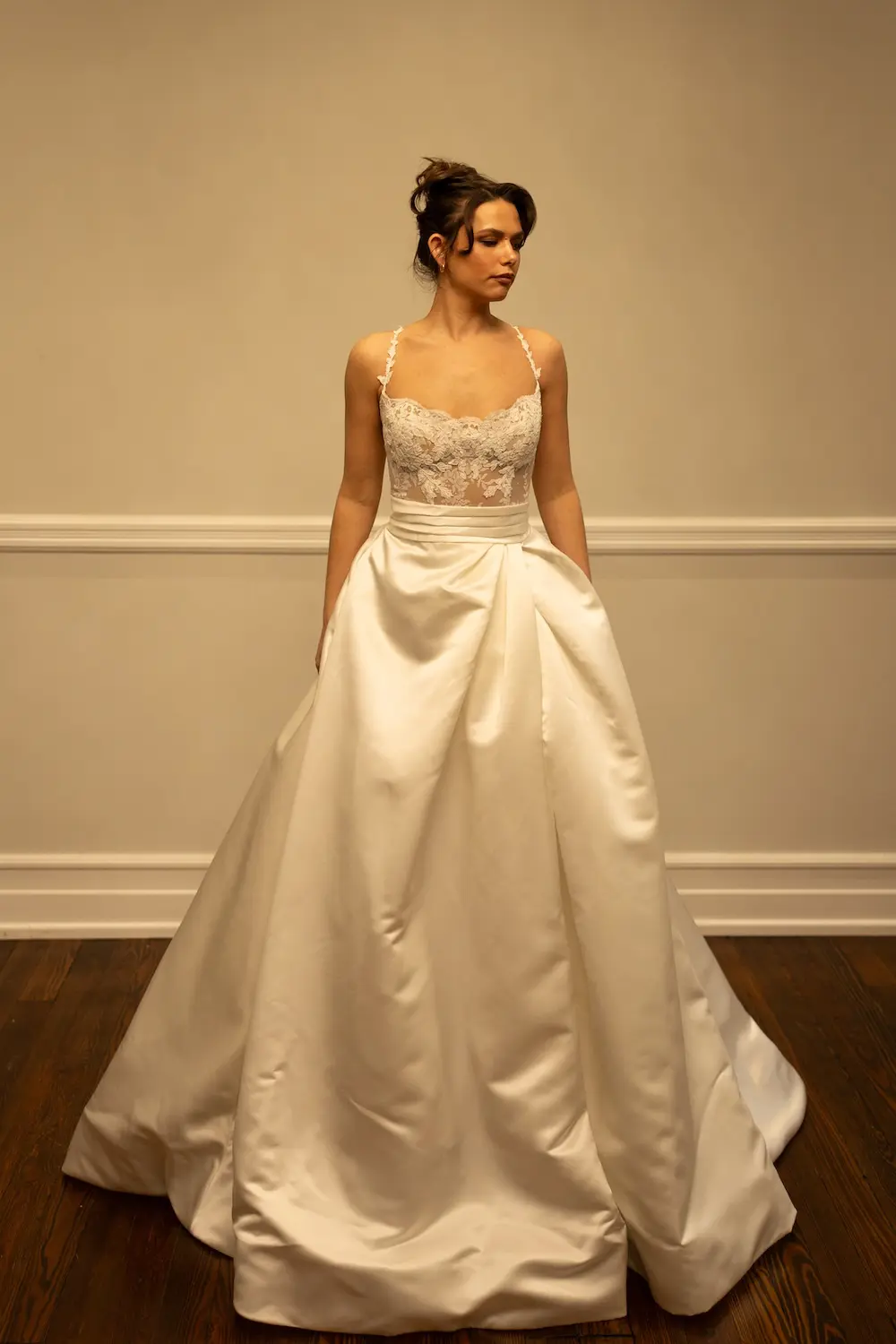 Morgan, an ivory and nude Sincerely Adorn bridal ball gown featuring a demi sheer lace bodice, a scoop neckline, and delicate spaghetti straps. Its voluminous satin skirt cascades to a chapel length train and includes button detailing.
