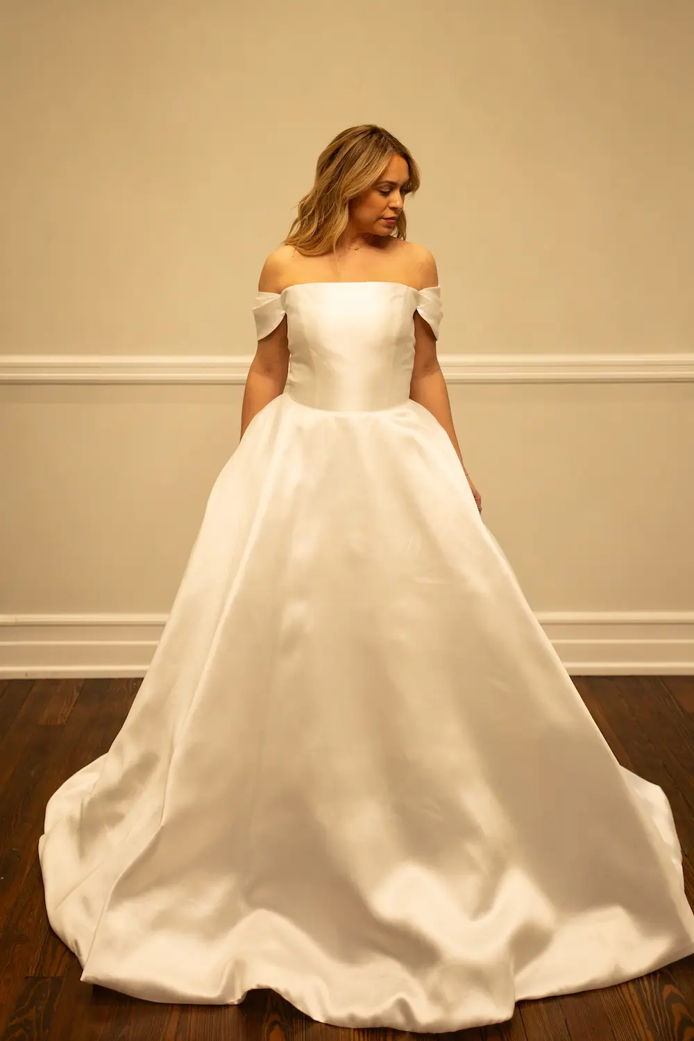 Kelly, an ivory bridal ball gown with an off-the-shoulder strapless design, crafted from lustrous Mikado fabric, featuring a voluminous skirt and a chapel train. A woman with blonde hair models the gown, looking gently downwards. Designer: Sincerely Adorn. Size: 12.