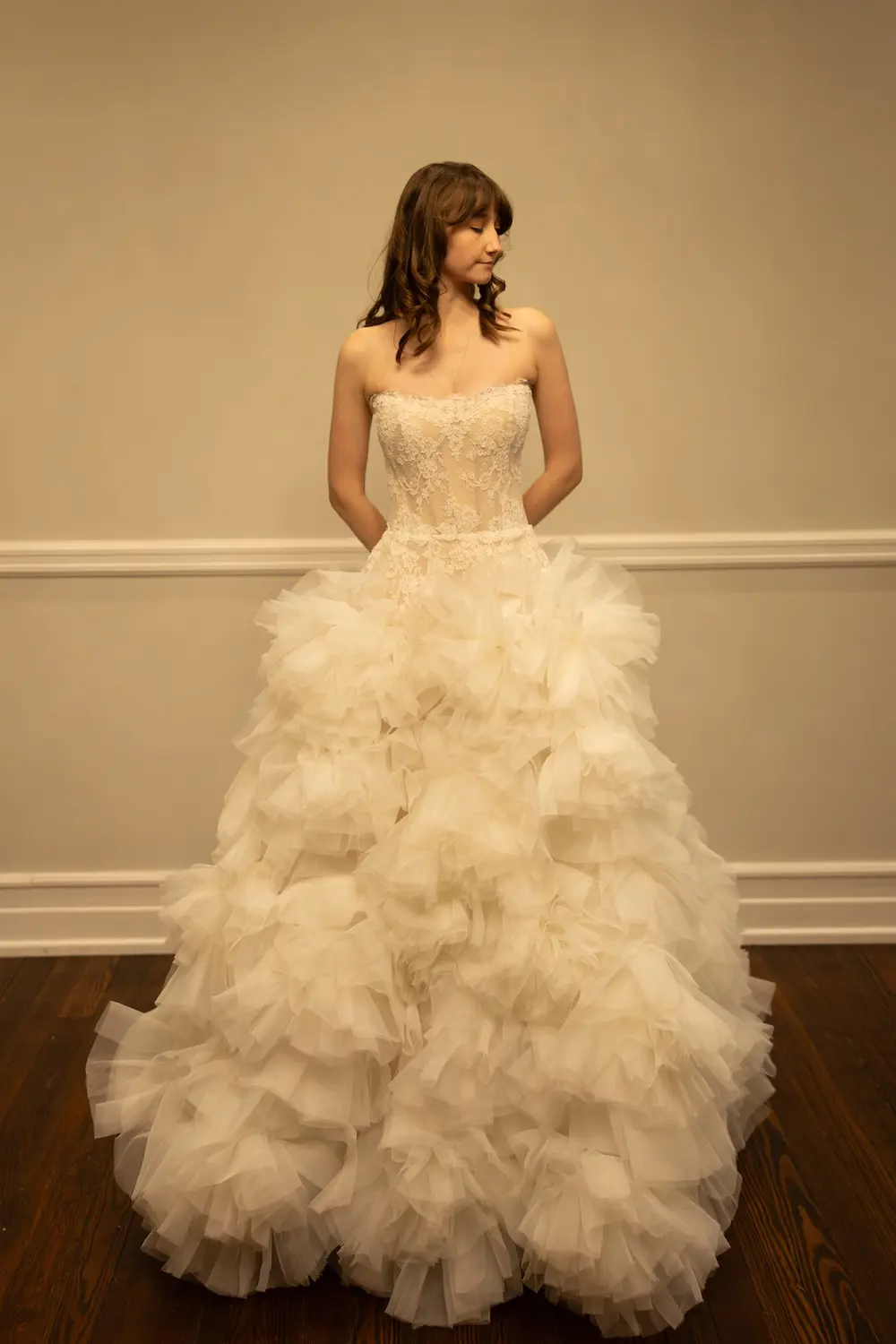 Audrey, an ivory bridal ball gown by Sincerely Adorn, featuring a strapless demi-sheer lace corset bodice and a dramatic, voluminous skirt made of cascading organza ruffles. A woman with shoulder-length wavy brown hair and bangs wears the gown, standing with her hands clasped behind her back and gazing softly to her right.
