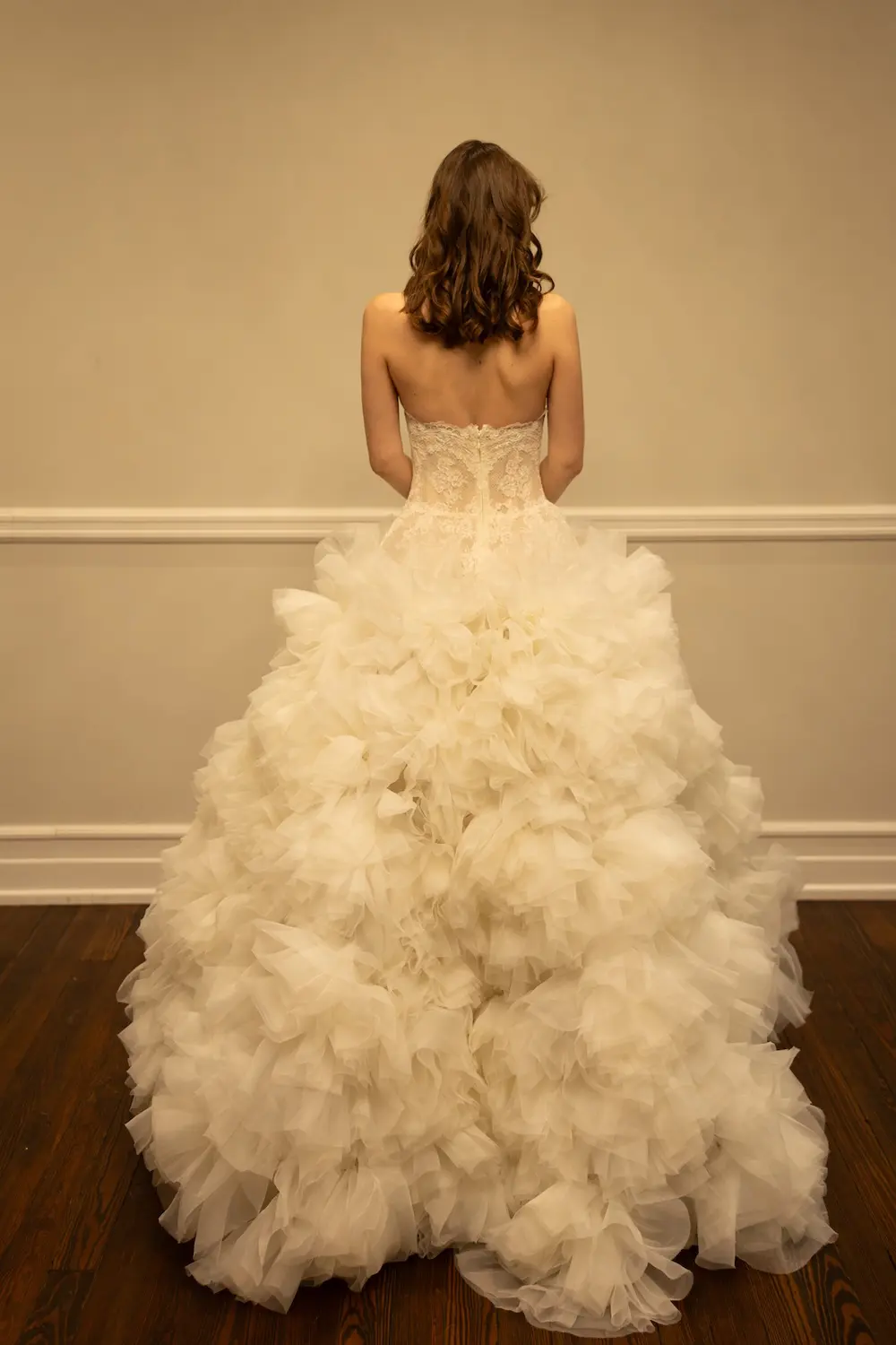 1771349861015-RH039-1 Audrey, an ivory ballgown wedding dress featuring a strapless, demi-sheer lace corset bodice and a dramatically voluminous organza skirt adorned with oversized sculptural ruffles, extending into a sweeping chapel-length train, seen from the back.